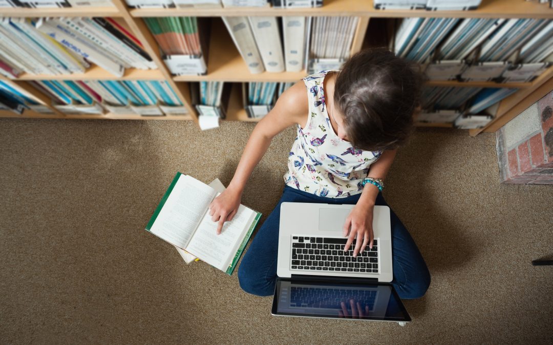 Student working on floor in library with laptop and books.