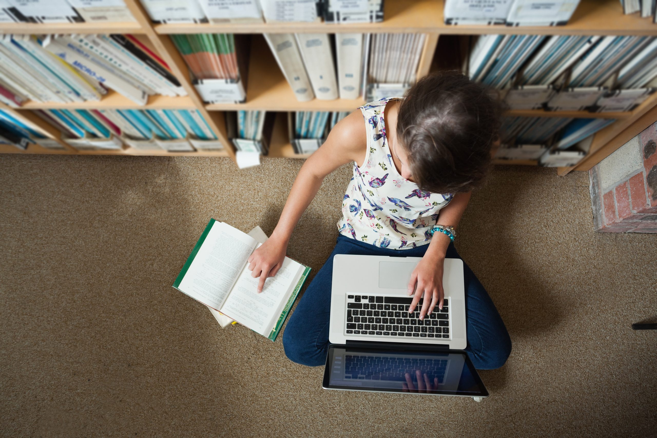 Student working on floor in library with laptop and books.