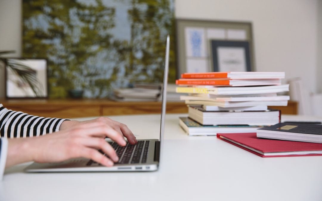 A person types on a laptop. A pile of books is behind the laptop.