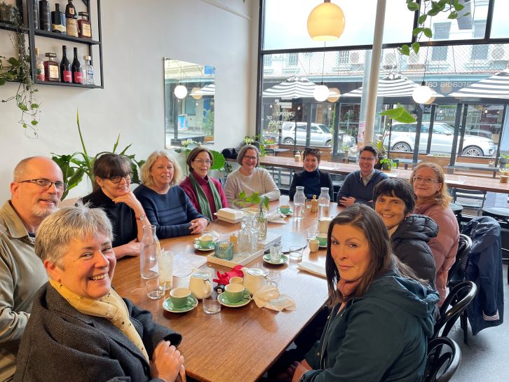 A group of people sit around a long table at a cafe. They are all looking towards the photographer and smiling. On the table are glasses and cups and saucers. Behind them is a large window looking out on the street.