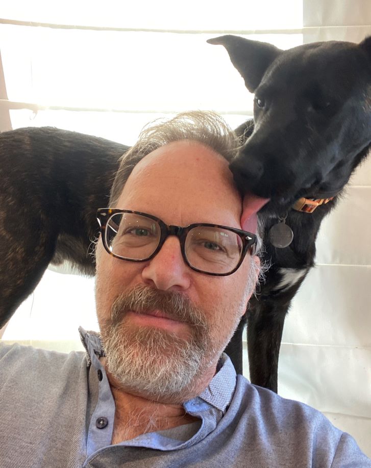 A man looks towards the camera. Standing behind him and looking over his shoulder is a black dog. The man's hair is grey white, and he has a beard in the same colour. He wear black rimmed glasses and a blue collared shirt. 
