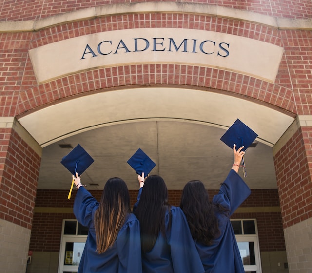 Three students dressed in navy blue graduation robes stand facing a brick building with a sign saying academics. They have their graduation caps raised as if to throw them into the air.