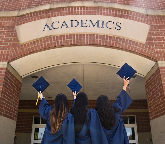 Three students dressed in navy blue graduation robes stand facing a brick building with a sign saying academics. They have their graduation caps raised as if to throw them into the air. 