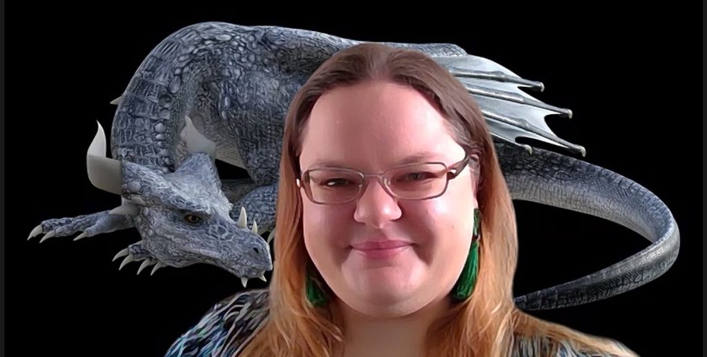 A woman looks to the camera, she's smiling and has long brown hair, green earrings and wears glasses. Behind her, is the image of a large grey dragon.