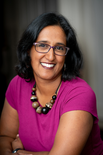 A portrait of a woman and shoulder length dark hair. She looks toward the camera and smiles. She wears glasses, a bright pink short sleeved top and a chunky necklace. 