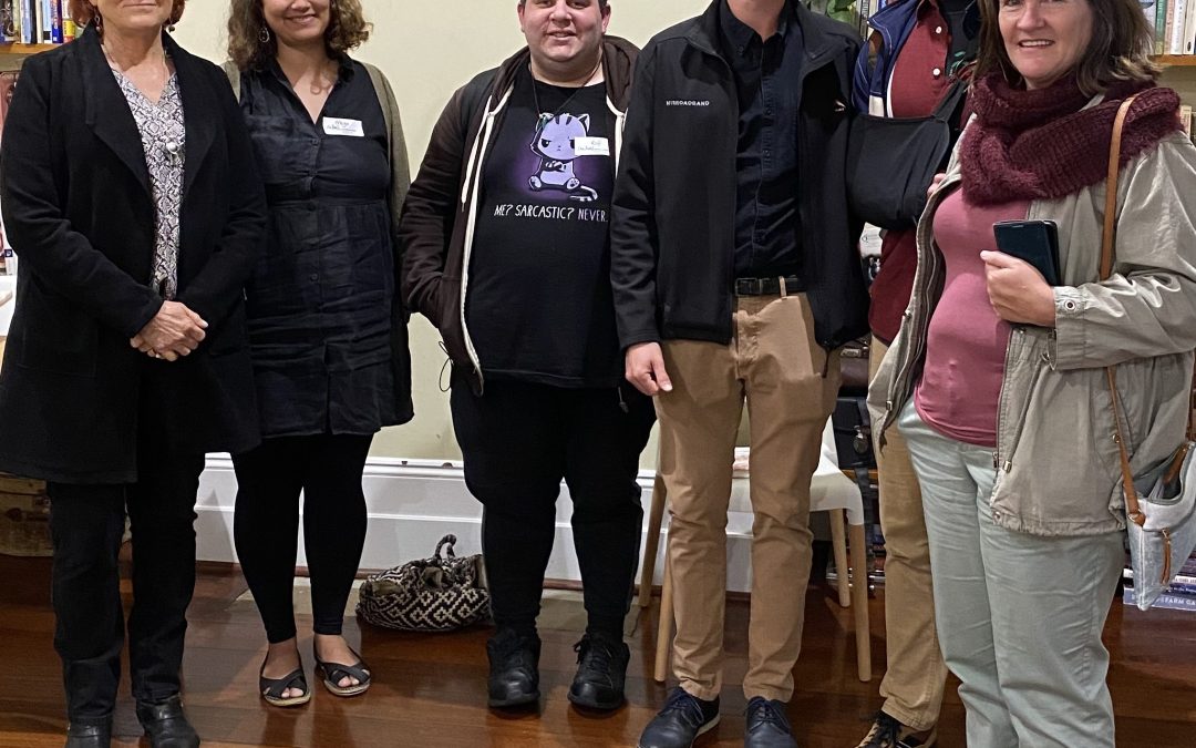 Six people stand together as a group, they look towards the camera and smile. Behind them are two large bookcases.