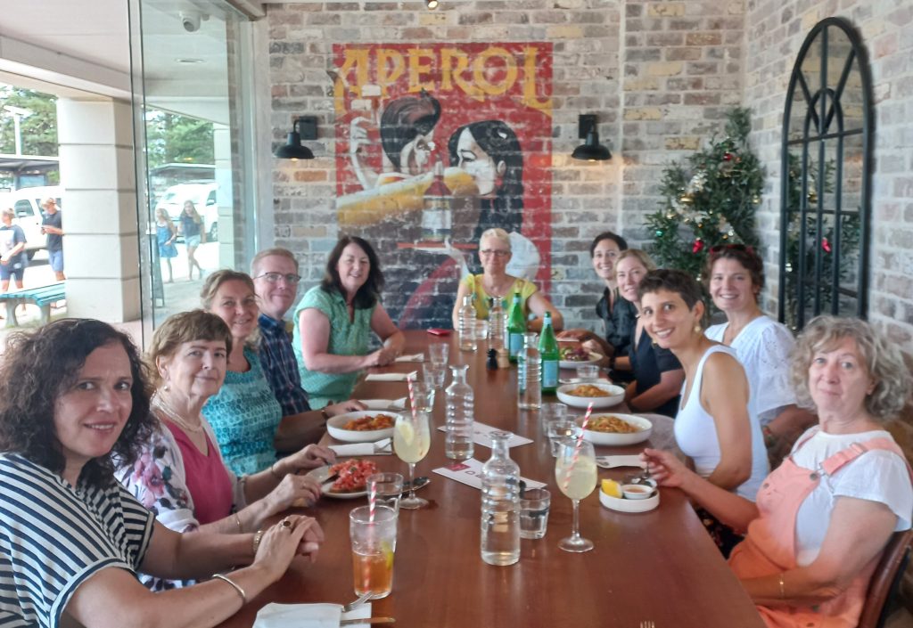 A group of people sit around a long table in a restaurant. There are plates of food, drinks and water jugs scattered across the table. It's daytime and light comes through a large glass window on the left hand side.