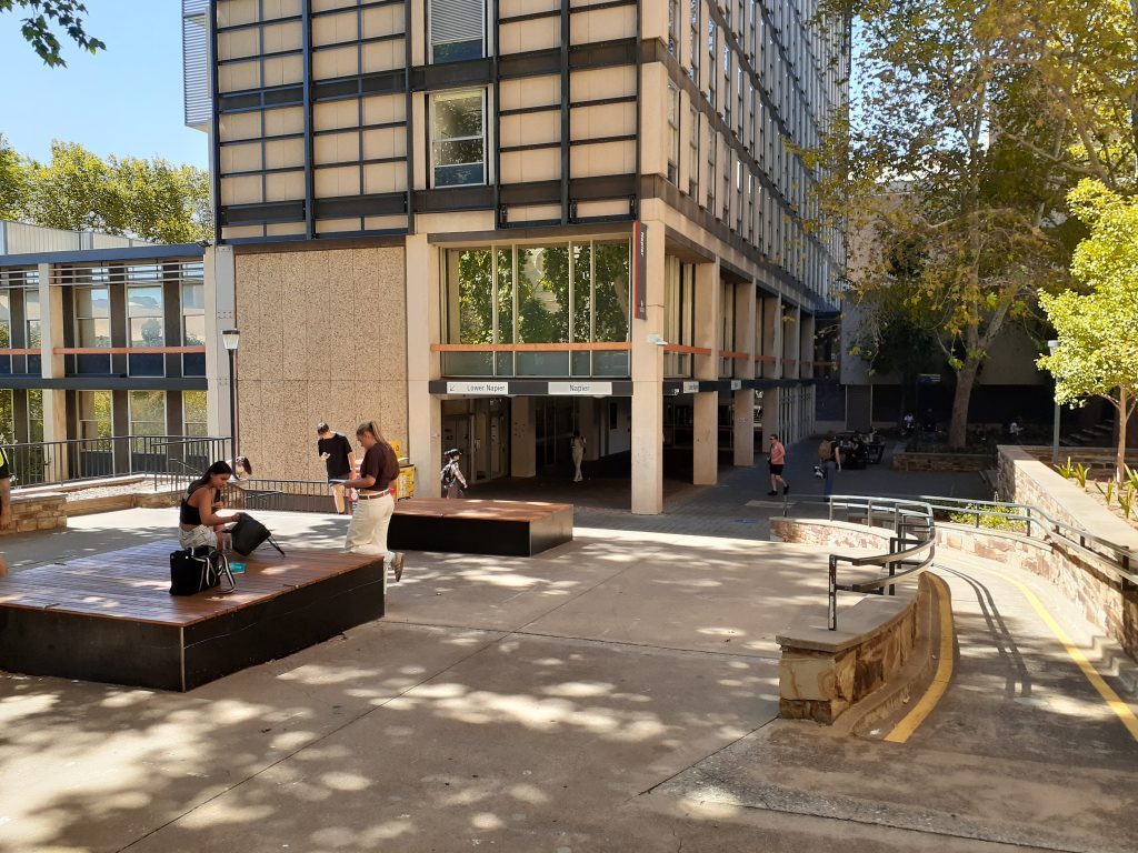 The external aspect of a building, including a view from the top of a ramp. A gradual ramp situated to the right of the stairs includes handrails along both sides and bright yellow vision-accessible strips painted along the ground on either side. The ramp's incline is gradual, and its path includes a slight bend.