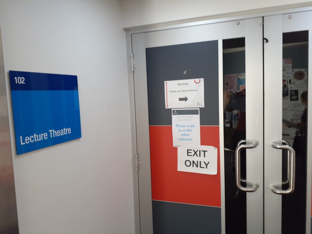 A set of double doors to a lecture theatre featuring D-shaped handles. These handles are positioned at an average height (level with middle hinge on the doors). The sign to the left reads 