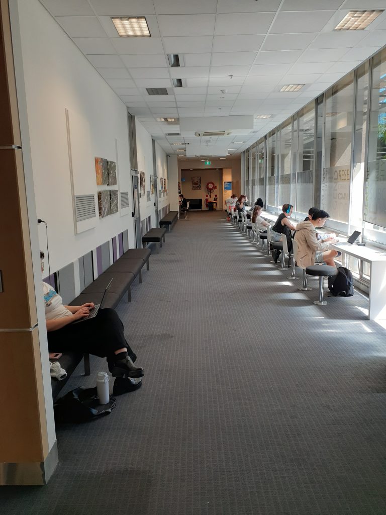 A hallway lined with low seating on the left side and desks on the right side. The desks include a combination of stools and chairs with backs. The desks face floor-to-ceiling windows.