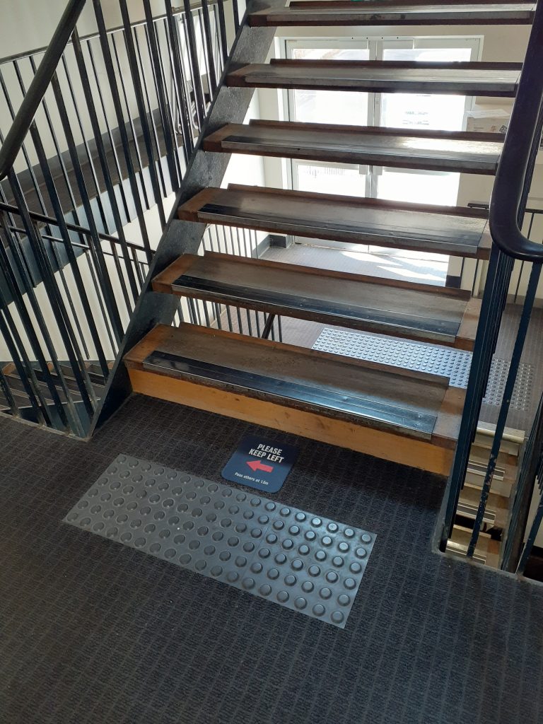 Image taken from a landing between flights of stairs. Two flights proceed downwards, and one flight is visible ascending upwards. The stairs are constructed of timber with metal nosing, and they include rails on both sides. Tactile indicators are located on the landings preceding the stairs in both directions. A sign prior to the stairs reads 