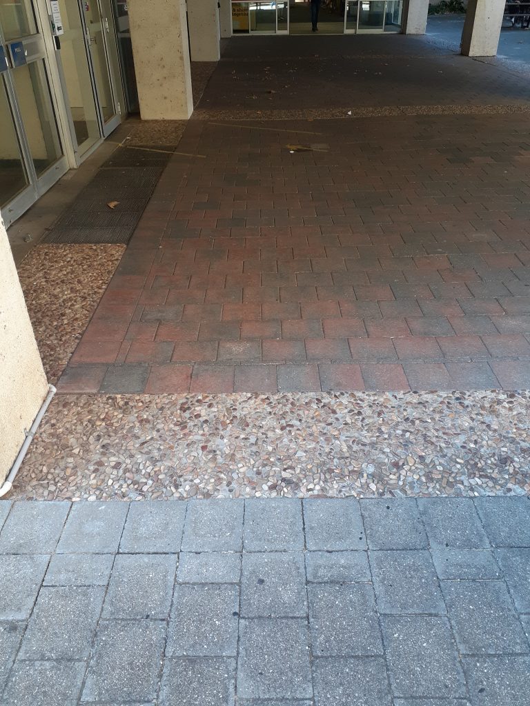An external pathway features different surface types, including pebbletex and brick. The transitions between these surface types are smooth, ensuring the pathway remains level access. Automatic sliding doors are situated directly ahead, and a set of push doors is visible to the left.