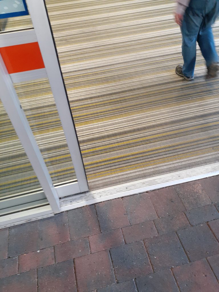 The transition between external and internal flooring at the threshold of an automatic sliding door. The external pathway is comprised of brick, and the internal flooring appears carpeted. A transition strip ensures this threshold remains level access.