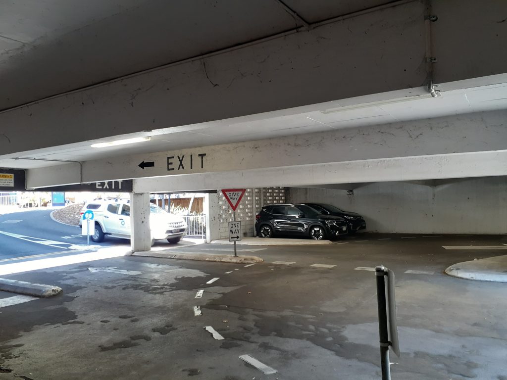 An undercover carpark entrance and exit. The cement flooring provides level acess, though no curb cuts are visible to support navigation of a mobility aid onto the paths or concrete islands pictured.